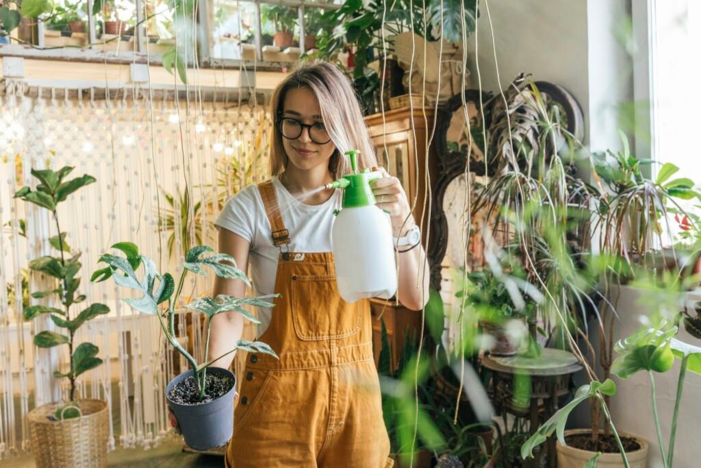 Mujer joven con gafas y overol amarillo sujetando una maceta con una planta de hojas verdes moteadas mientras sostiene un pulverizador blanco con boquilla verde. Está en un espacio interior rodeada de plantas, decoraciones y una cortina de cuerdas.