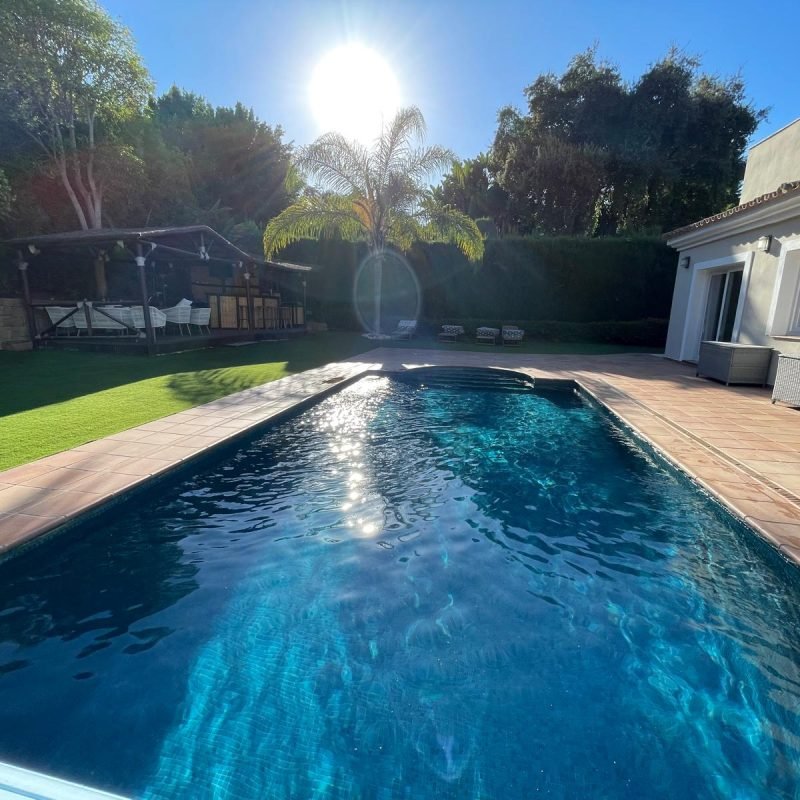 WhatsAppImage20241021at11101761729503934 Piscina con agua azul clara reflejando la luz del sol, rodeada de una terraza pavimentada. Al fondo hay una estructura de madera techada y varias tumbonas, todo rodeado de vegetación.